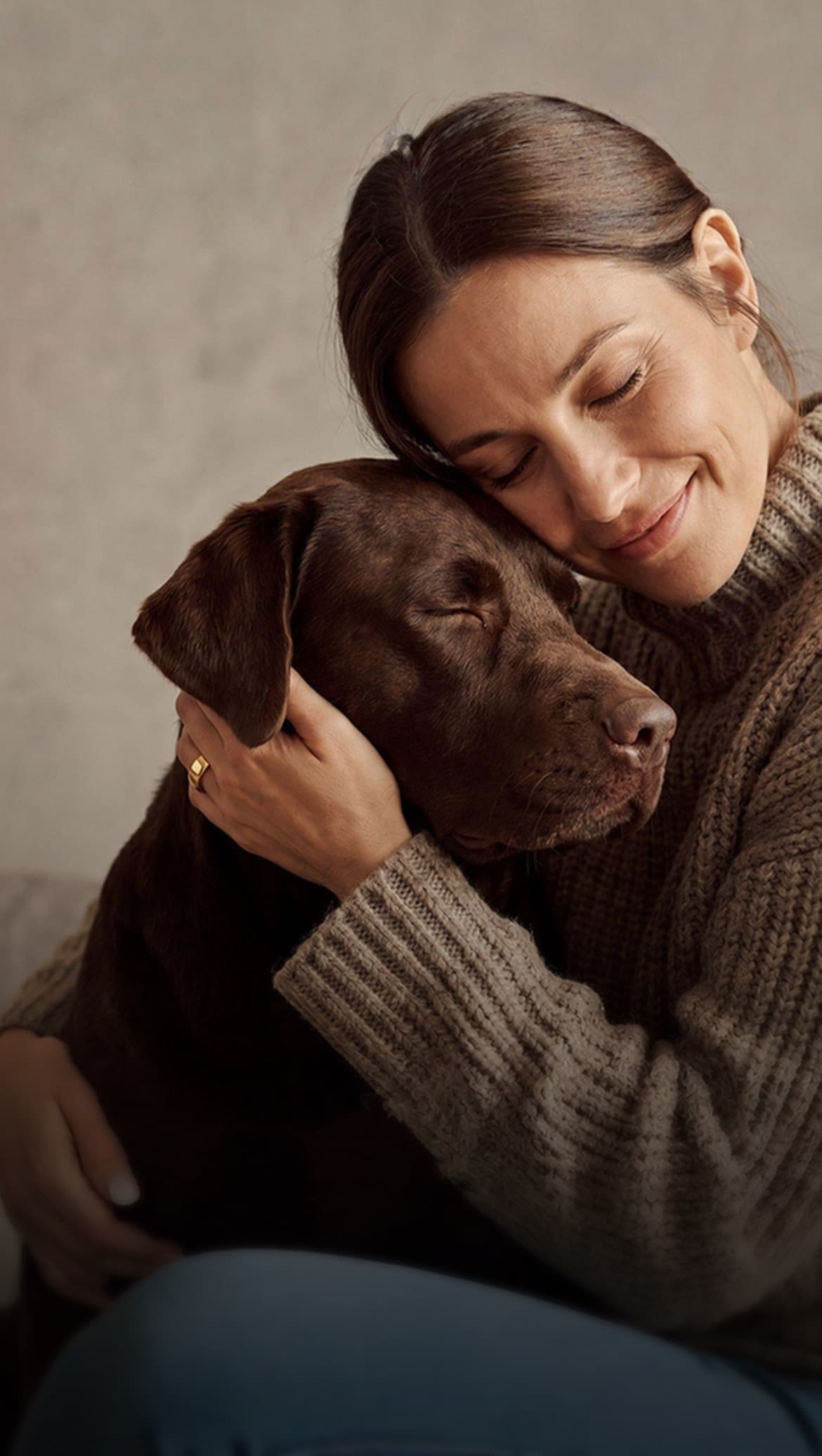 A woman rests her cheek on her chocolate Labrador, both with their eyes closed.