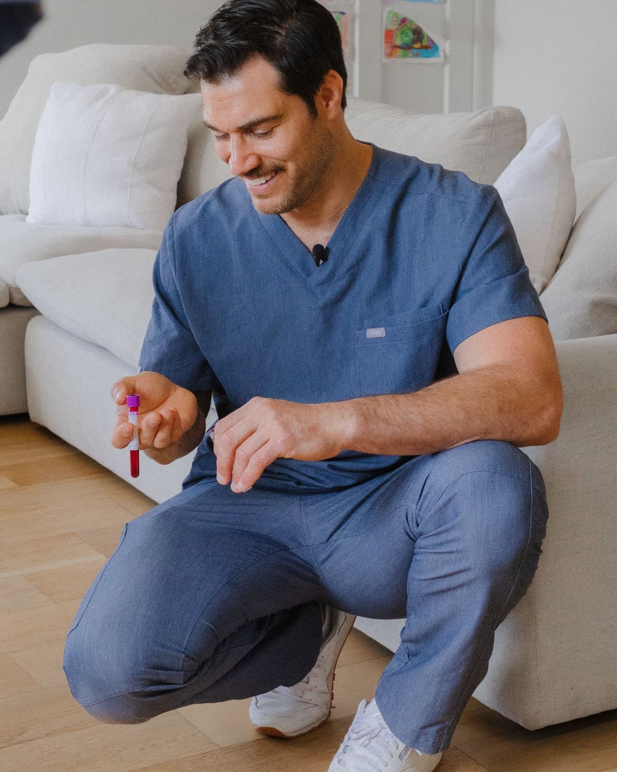 Dr. Evan Antin, DVM, in blue scrubs kneels on a wood floor, smiling as he holds a blood-sample vial.